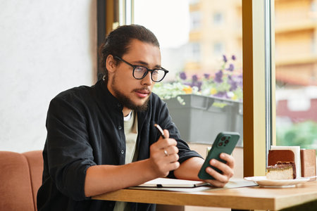 A young man with a beard is focused on his smartphone and notes in a cafe.の写真素材