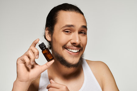 Handsome young man with a beard holds up a bottle of essential oil, radiating joy and positivity.の写真素材
