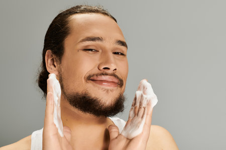 Bearded young man happily cleansing his face with a foamy wash, showcasing confidence and care.の写真素材