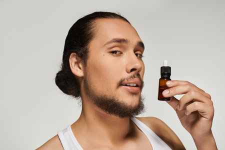 Young bearded man smiles while holding a bottle of skincare serum in a bright setting.の写真素材