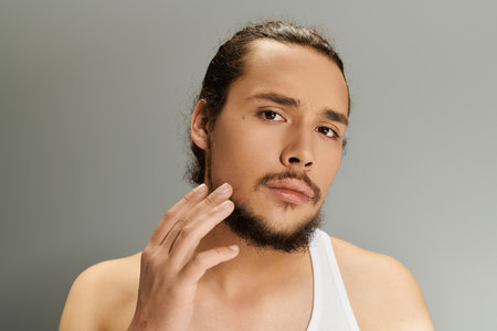 A young man with a beard touches his chin, exuding charm and confidence in a soft lit setting.の写真素材