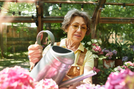 A senior beautiful woman gently waters bright flowers in her lush garden during a sunny day.の写真素材