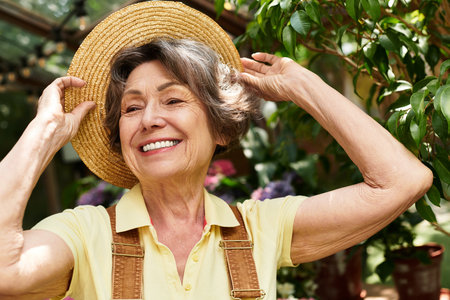 A senior woman enjoys the beauty of her colorful garden, smiling as she adjusts her sunhat.