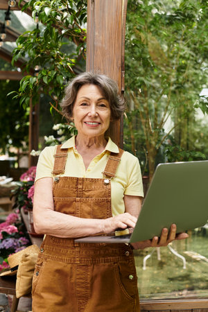 In a vibrant garden, a joyful senior woman shares her love for plants while using a laptop.の写真素材