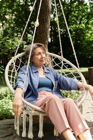 A beautiful senior woman relaxes in a cozy swing, surrounded by lush greenery and vibrant flowers.の写真素材