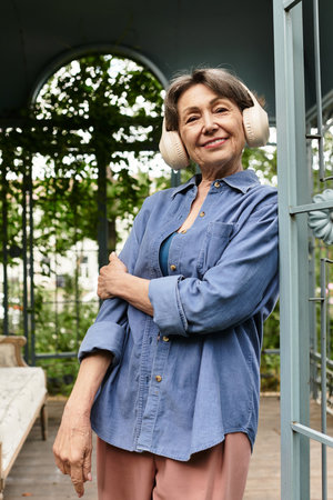 Beautiful senior woman relaxes in a serene garden, wearing headphones and smiling warmly.
