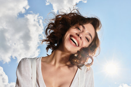 A young woman with wavy brown hair smiles joyfully under a clear blue sky on a summer day.の写真素材