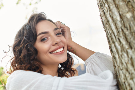 A young woman with wavy brown hair smiles brightly while outdoors in a sunny park.の写真素材