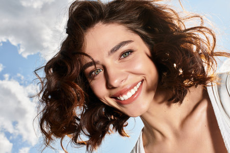 A pretty young woman with wavy brown hair smiles joyfully under a vibrant summer sky outdoors.の写真素材
