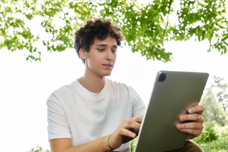 A young man with curly hair relaxes in a green park, engaging with his tablet under the warm sun.の写真素材