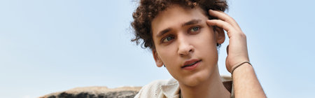 A young man with curly hair relaxes in a lively park, showcasing his fashionable summer look, banner