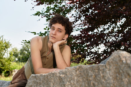 A young man with curly hair relaxes in a green park, showcasing his trendy summer outfit.