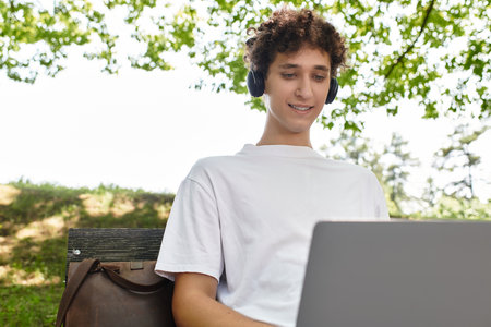 Curly-haired youth relaxes outdoors, focused on his laptop in a sunny green park.の写真素材