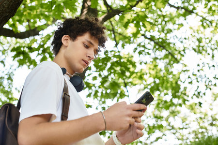 Curly-haired youth relaxes outdoors in a vibrant park while checking his phone.の写真素材