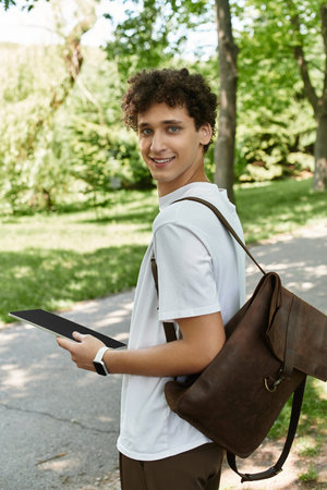 A young man with curly hair relaxes in a vibrant summer park, engaging with a tablet.