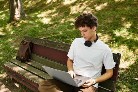 A young man with curly hair enjoys his time outdoors, working on a laptop in a green park.の写真素材