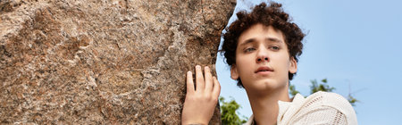 A young man with curly hair enjoys a sunny day in a green park, showcasing his unique fashion sense,...