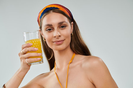 A pretty young woman with tan skin and a headscarf poses, holding a glass of juice in summer attire.の写真素材