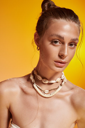 A confident young woman poses in a summer outfit and seashell necklace in a studio.