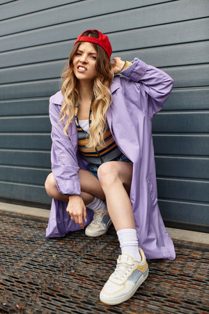 Smiling young woman in purple coat and cap poses confidently in a playful summer aesthetic.