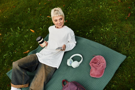A young woman in a stylish outfit relaxes in a park, sipping coffee and enjoying her phone.