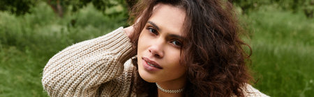 Young woman with curly hair poses in a chic summer outfit while surrounded by lush greenery, banner