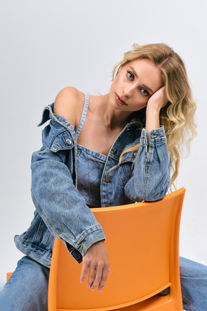 Blonde woman elegantly shares her fashion flair while resting on a bright chair indoorsの写真素材