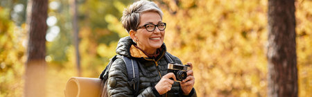 A joyful senior woman captures beautiful moments while hiking amidst lush autumn forest scenery.