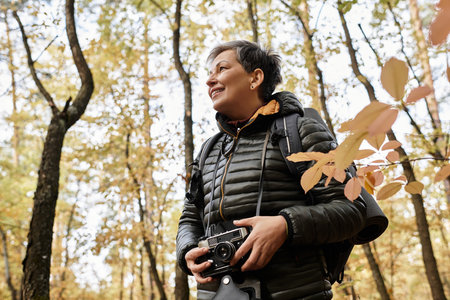 Senior woman delights in nature as she hikes through a vibrant autumn forest, camera in hand.