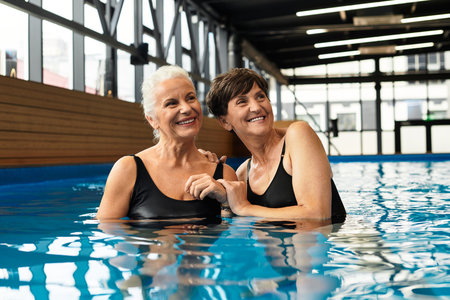 Two beautiful mature women embrace joyfully while surrounded by calming water in a wellness center.