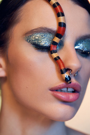 A young woman poses beautifully with a vibrant snake coiled on her face, showcasing makeup.