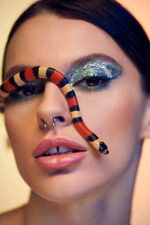 A stunning young woman poses with bold makeup and a colorful snake on her face.