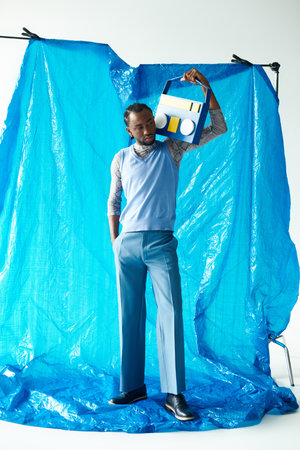 A young man with a charming smile stands tall in stylish attire against a blue backdrop.
