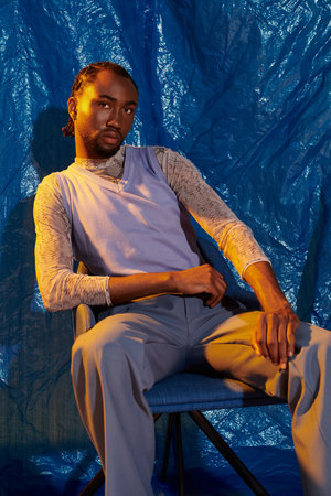A charming young man poses confidently on a chair against a vivid blue background.