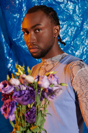A striking young man with a confident expression stands holding a colorful bouquet of flowers.