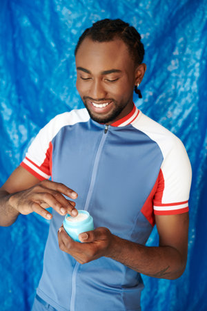 Young man smiles joyfully while applying skincare cream against a bright blue backdrop.の写真素材