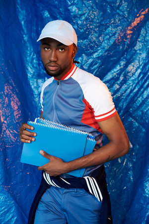 A stylish young man confidently holds blue notebooks against a vibrant backdrop.