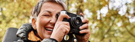 Senior woman joyfully takes pictures with her camera during a hike in a vibrant forest.の写真素材