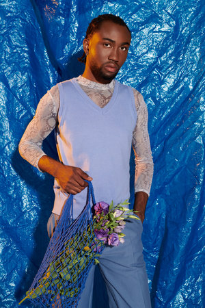 Handsome young man poses confidently with a net bag of flowers in front of a striking blue backdrop.