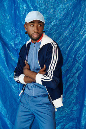 A stylish young man stands with arms crossed, exuding confidence against a striking blue background.