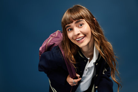 A joyful young woman with long hair beams as she holds her backpack, embracing her studious spirit.