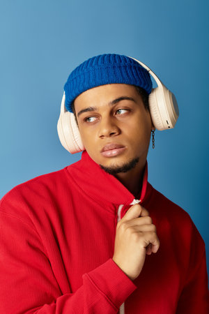Stylish young Black man in a red sweatshirt and blue beanie stands confidently, looking away.の写真素材