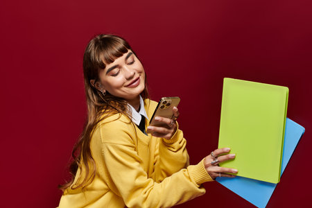 Smiling young woman in a yellow sweater holds colorful notebooks and a smartphone while studying.