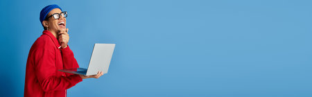 A stylish young Black man in a beanie and glasses smiles happily with a laptop, bannerの写真素材