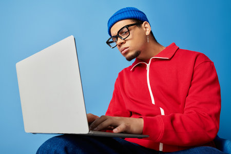 A stylish young man in a beanie focuses intently on his laptop while seated comfortably.