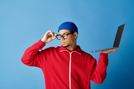 Stylish young african american man in beanie poses confidently with a laptop and blue backdrop.