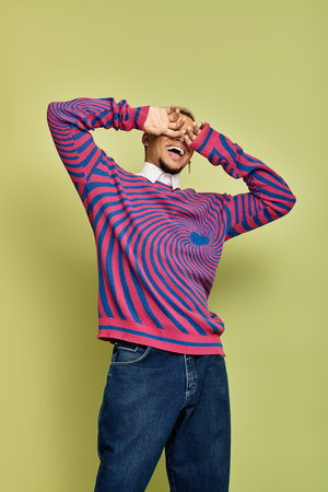 A fashionable young man joyfully engages with the camera, showcasing a colorful striped sweatshirt.