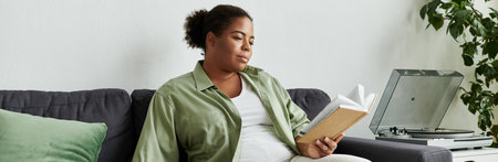 An African American woman enjoys a quiet afternoon at home reading while lounging on the couch,...