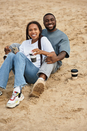 Loving couple sits closely on sandy beach, sharing smiles and warm drinks while enjoying their time.