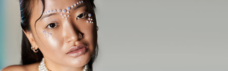 A young Asian woman showcases unique makeup artistry with pearls on her face in a studio, banner